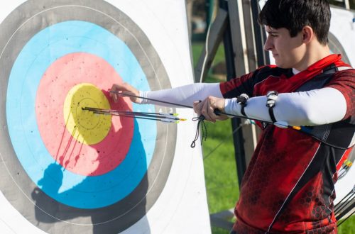 Focus on man aiming with bow and arrows at target during outdoor archery practice.