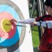 Focus on man aiming with bow and arrows at target during outdoor archery practice.